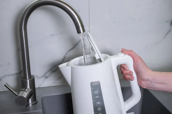 A woman draws water into an electric kettle in the background of the kitchen. It's time for breakfast and tea. Modern electric kettle. Kettle for boiling water.