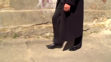 Siyah uzun paltolu genç bir adam eski bir binanın yanındaki asfaltta yürür ve koşar.
