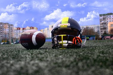 Bir Amerikan futbol topu ve miğferi bulutlu gökyüzüne karşı yeşil çimlerin üzerinde duruyor.