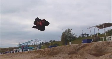 Bir adam deniz kıyısında, bulutlu bir gökyüzüne karşı ağır çekimde akrobatik bir vuruş sergiliyor.