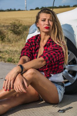 Siyah ve kırmızı kareli gömlekli, uzun saçlı güzel bir kız bir arabanın ön tekerleğinin yanında oturuyor.