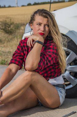 Siyah ve kırmızı kareli gömlekli, uzun saçlı ve kısa şortlu güzel bir kız bir arabanın ön tekerleğinin yanında oturuyor.