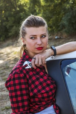 Kareli tişörtlü, elinde İngiliz anahtarıyla güzel bir kız arabanın kapısına dayanıyor.