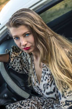 Mavi gözlü, uzun saçlı, güzel bir kız beyaz bir arabanın kapısından dışarı bakıyor.