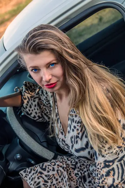 Mavi gözlü, güzel, benekli, uzun saçlı, güzel bir kız. Beyaz bir arabanın kapısından dışarı bakıyor.