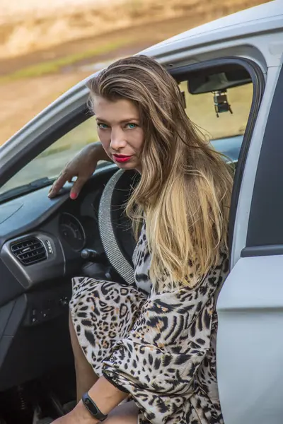 Mavi gözlü, güzel, uzun elbiseli, uzun saçlı güzel bir kız beyaz bir arabanın kapısından dışarı bakıyor.