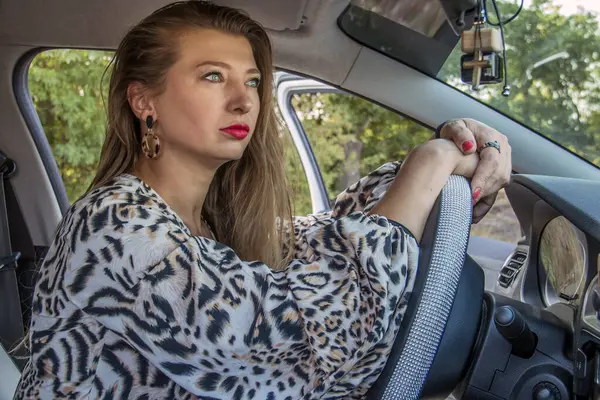 Güzel, açık renk elbiseli güzel bir kız direksiyonun arkasında bir arabada oturuyor.