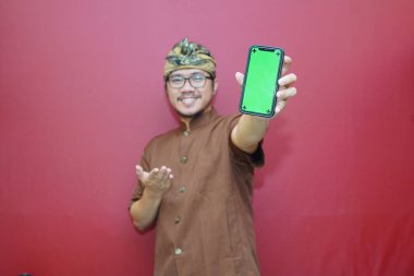 Udeng giyen heyecanlı Balili adam ve kırmızı arka planla izole edilmiş akıllı telefon ekranını gösteren geleneksel kıyafetler. Modelleme için boş alanı olan Gadget.