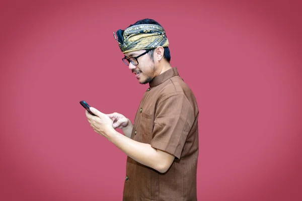Mutlu bir Balili adamın portresi gülümsüyor ve elinde udeng ve kırmızı arka planla izole edilmiş geleneksel kıyafetlerle akıllı telefonunu tutuyor..