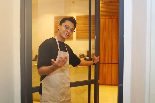Portrait of smiling owner standing and opening his restaurant door. The chef or waiter stands in front of the coffee shop and looks at the camera with an inviting gesture.