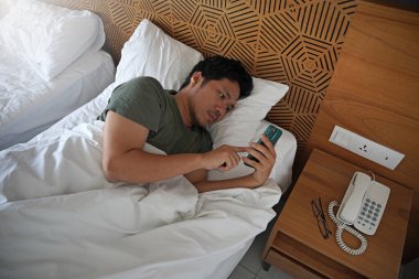 Gülümseyen Asyalı adam, akıllı telefondan sosyal medyayı kontrol ederken rahat bir yatakta yatarken uyanıyor, mutlu adam sabah uyandığında cep telefonu kullanıyor ve hoş bir mesaj veya mesaj alıyor.