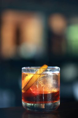 Klasik bir kokteylin yakın plan çekimi, muhtemelen bir Negroni ya da benzer bir içki, düşük bardakta. İçkinin kehribar kırmızısı renkli ve portakal kabuğuyla süslenmiş..