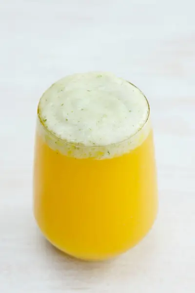 Stock image A close-up shot of a vibrant yellow smoothie or juice in a clear glass, topped with a frothy layer. The drink appears fresh and inviting, with tiny green flecks in the froth.