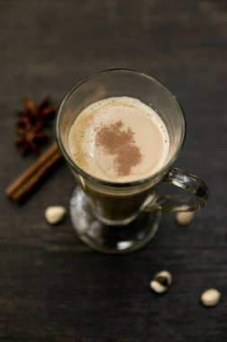 A close-up shot of a spiced chai latte in a clear glass mug, topped with a sprinkle of cinnamon. The warm beverage is set against a dark, textured background, with star anise, cinnamon sticks. clipart