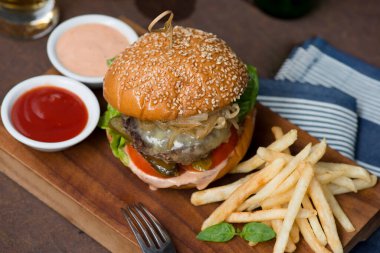 Tahta bir tahtanın üzerinde servis edilen gurme bir çizburgerin yakın plan görüntüsü. Hamburgerde susamlı çörek, erimiş peynir, karamelize soğan, domates, marul ve turşu yer alıyor.. 