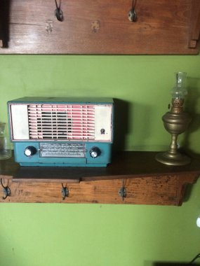 Görüntü, antika bir radyo ve yeşil bir duvara dayalı ahşap bir rafa yerleştirilmiş bir gaz lambasını gösteriyor. Radyo, mavi bir vücudu ve metal ızgarası olan klasik bir model..