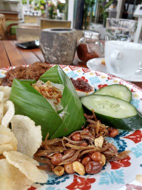 Görüntü canlı ve iştah açıcı bir Endonezya yemeği sergiliyor. Muz yaprağına sarılı beyaz pirinç, çıtır krakerler, baharatlı ançüez, fıstık ve dilimlenmiş salatalıkla birlikte orta kısımdır.