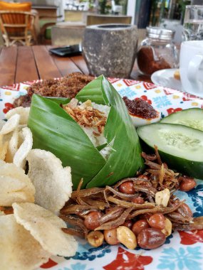 Görüntü canlı ve iştah açıcı bir Endonezya yemeği sergiliyor. Muz yaprağına sarılı beyaz pirinç, çıtır krakerler, baharatlı ançüez, fıstık ve dilimlenmiş salatalıkla birlikte orta kısımdır.