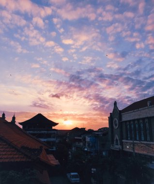 Görüntü, şehir siluetinin üzerinde renkli bir günbatımını gösteriyor. Gökyüzü turuncu, pembe ve mor renklerle parıldıyor, ve şehir binaları canlı arkaplana karşı bir siluete bürünüyor.