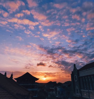 Görüntü, şehir siluetinin üzerinde renkli bir günbatımını gösteriyor. Gökyüzü turuncu, pembe ve mor renklerle parıldıyor, ve şehir binaları canlı arkaplana karşı bir siluete bürünüyor.