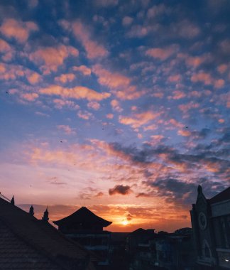 Görüntü, şehir siluetinin üzerinde renkli bir günbatımını gösteriyor. Gökyüzü turuncu, pembe ve mor renklerle parıldıyor, ve şehir binaları canlı arkaplana karşı bir siluete bürünüyor.