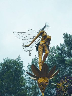 Malang, 20 Haziran 2018. Görüntü, çiçeğin tepesinde duran kanatlı bir peri heykelini gösteriyor. Çiçeğin büyük pembe yaprakları ve sarı bir merkezi vardır. Heykel dışarıda bir bahçede bulunuyor..