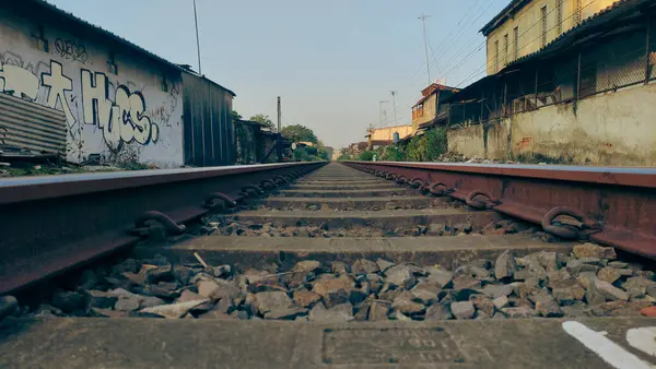 stock image The image depicts a straight stretch of railway tracks extending into the distance. The tracks are flanked by a sparse arrangement of buildings on either side.