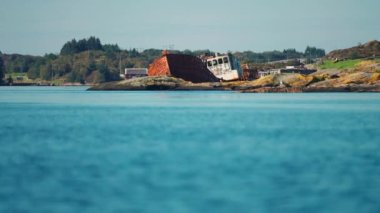 A rusty shipwreck on the rocky shore of the Atlantic. Several small houses are scattered along the shores. High-quality 4k footage