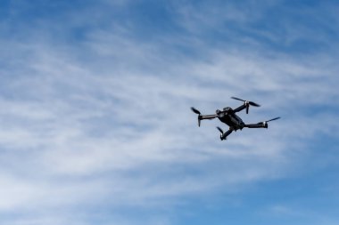 Çerçevenin sağ yarısında, ince beyaz bulutları olan mavi bir gökyüzüne karşı kurulmuş bir dron var..