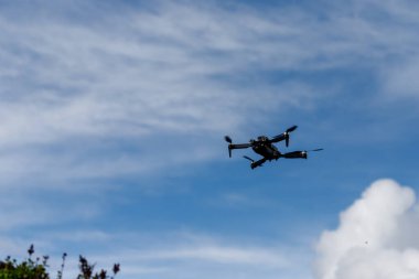 Siyah bir dron havada, dağınık beyaz bulutlu mavi gökyüzünün arkaplanına doğru süzülüyor. Sol alt köşede bazı bitkiler görünür..