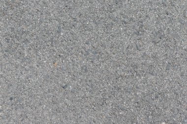 Gri asfalt yakın çekim. Yüzeyde küçük çakıl ve çakıl taşları gömülüdür, desenli ve dengesiz bir görünüm yaratır..
