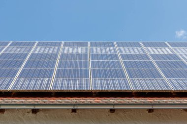 Mavi gökyüzünün altındaki güneş panelleriyle kaplı bir çatı. Paneller kiremitli bir çatının üzerinde, bir oluk ve aşağı görünür beyaz bir duvar ile.