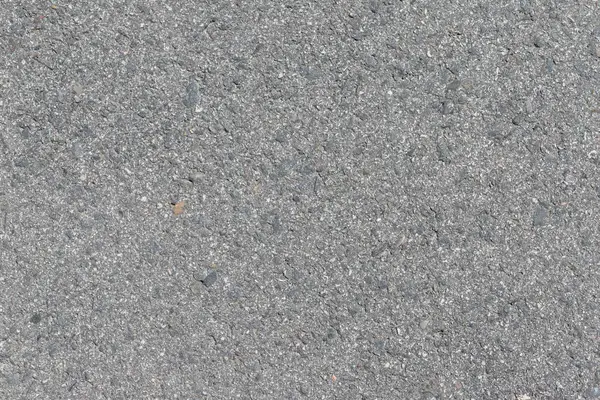 Gri asfalt yakın çekim. Yüzeyde küçük çakıl ve çakıl taşları gömülüdür, desenli ve dengesiz bir görünüm yaratır..