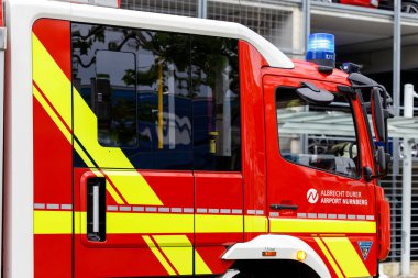 Nuremberg, Almanya - 25 Mayıs 2025: Sarı çizgili kırmızı ve beyaz bir itfaiye aracının yan ve önü, pencereler, mavi ışık ve havaalanı adı.