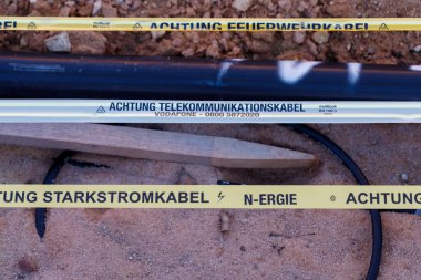 Nuremberg, Almanya 2025 Mart 02: Uyarı bantlarıyla işaretlenmiş gömülü kablo ve elektrik hatlarının zemin görüntüsü. Farklı uyarı bantları ve bir parça ahşap işaret aracı konumu.