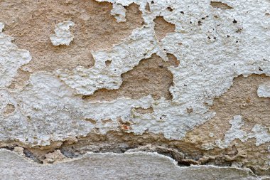 Beyaz boyanın soyulduğu bir duvara yakın plan, altta yatan kahverengi ve gri malzeme katmanlarını ortaya çıkarıyor. Doku pürüzlü ve yaşlanmış..