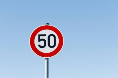 Metal bir direğin üzerinde açık mavi gökyüzüne karşı beyaz, kırmızı ve siyah 50 hız sınırı tabelası. İşaret solda, gökyüzü sağda..