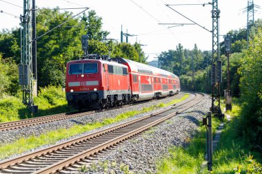 Güneşli bir günde yeşillikle çevrili raylar boyunca birden fazla vagon kıvrımlı kırmızı bir DB treni. Tepedeki güç hatları görülebilir..