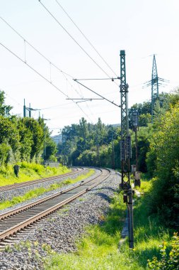 Uzağa doğru kıvrılan tren raylarının manzarası, ağaçlarla kaplı, ve yukarıdaki elektrik hatları. Metal kutuplar ve elektrik ekipmanları görünür..