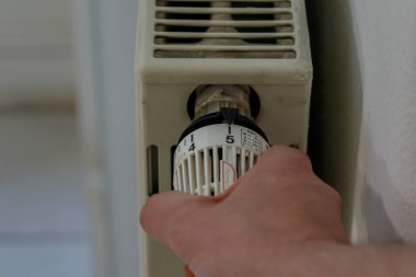 Radyatörün termostat vanasını ayarlayan bir elin yakın çekimi. Termostat 5 'e ayarlı. Radyatör beyaz değil, beyaz bir duvara monte edilmiş..