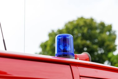 Mavi bir acil durum ışığı ve kırmızı hoparlörlü, arka planda bulanık bir ağaç ve beyaz gökyüzü olan kırmızı bir aracın yakın plan görüntüsü..