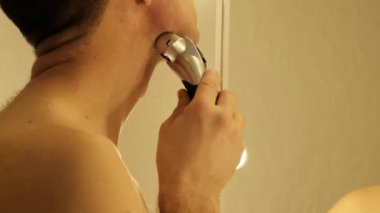 Bir adam aynanın önünde dikilirken elektrikli jiletle tıraş olur. Yüksek kalite 4k görüntü