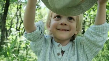 Üç yaşında gömlekli bir çocuk şapkasını sallıyor ve gülümsüyor. Arka planda doğa var. Yüksek kalite 4k görüntü