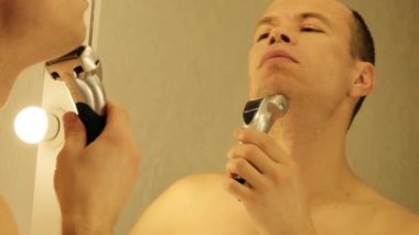 Bir adam aynanın önünde dikilirken elektrikli jiletle tıraş olur. Yüksek kalite 4k görüntü