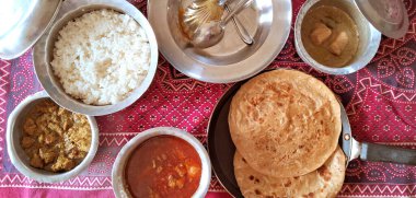 Kırmızı desenli masa örtüsü üzerinde pirinç, köri ve yassı ekmek gibi çeşitli yemekler düzenlenir. Paslanmaz çelik kaseler ve tabaklar yiyecekleri tutarak geleneksel bir yemek ortamı oluşturuyor..