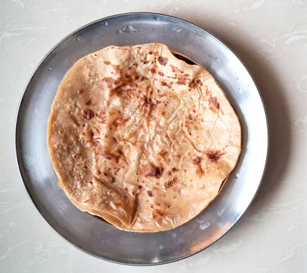 Yeni pişmiş bir paratha paslanmaz çelik bir plakaya yerleştirilir ve altın kahverengi benekleri sergilenir. Arkaplanda açık renkli mermer bir yüzey var..