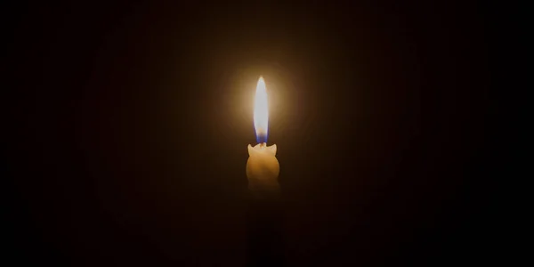 A single burning candle flame or light is glowing on a spiral white candle on black or dark background on table in church for Christmas, funeral or memorial service.