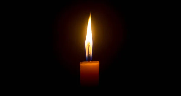 A single burning candle flame or light is glowing on an orange candle on black or dark background on table in church for Christmas, funeral or memorial service with copy space.