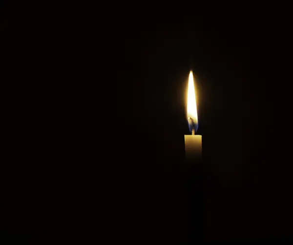 A single burning candle flame or light is glowing on small orange candle on black or dark background on table in church for Christmas, funeral or memorial service with copy space