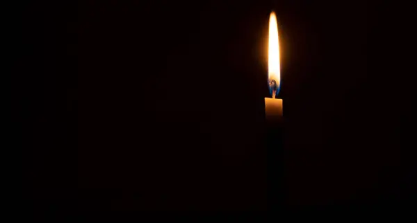 A single burning candle flame or light is glowing on an orange candle on black or dark background on table in church for Christmas, funeral or memorial service with copy space